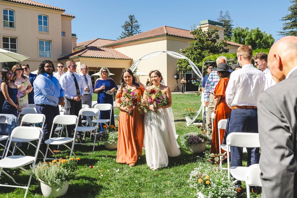 Bride walks down outdoor wedding aisle with bridesmaid in rust dress, guests seated on white chairs, elegant venue backdrop.