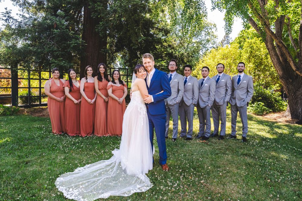 Bride and groom pose with bridesmaids in rust dresses and groomsmen in gray suits outdoors.