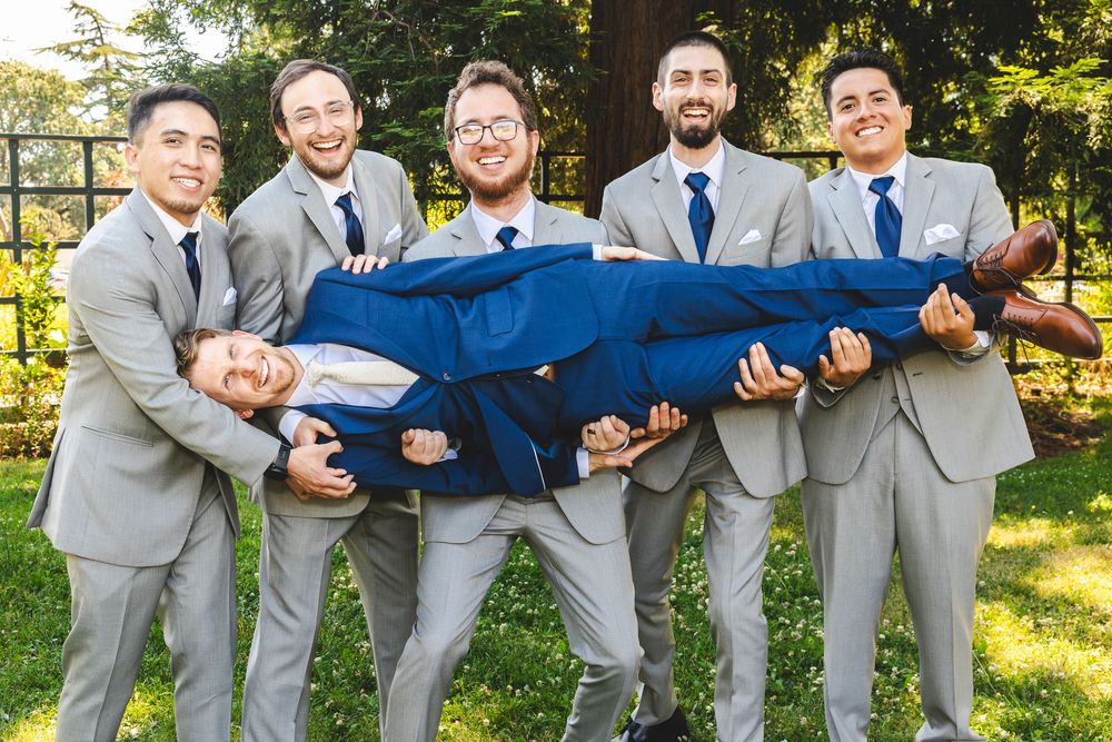 Groom in blue suit lifted by four groomsmen in grey suits, laughing outdoors at wedding reception.