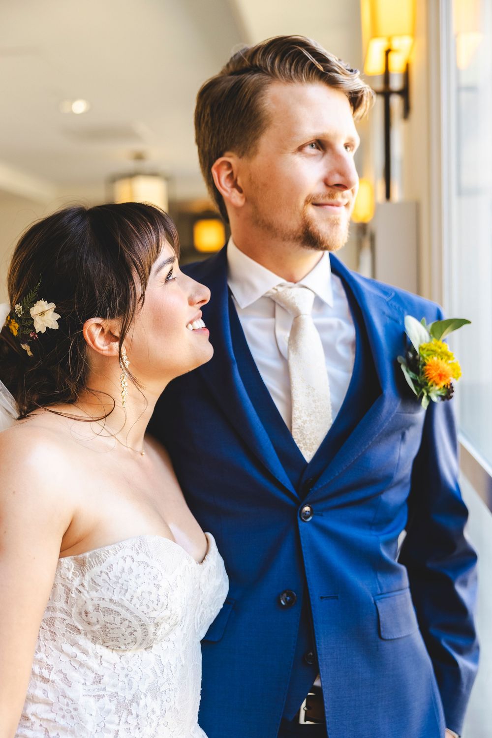 Bride in lace gown gazes lovingly at groom in blue suit with boutonniere near sunlit window.