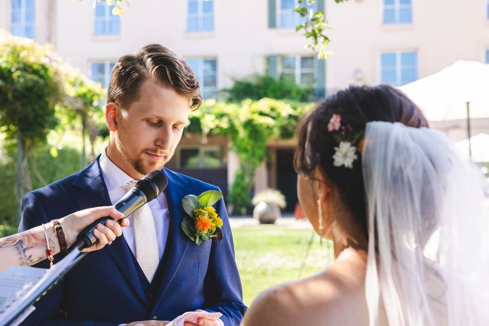 Groom in blue suit reading wedding vows into microphone beside bride in white veil with floral hair accessories outdoors.