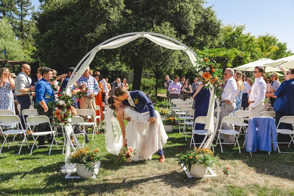 A couple shares a kiss under a white arch at an outdoor wedding ceremony with guests seated.