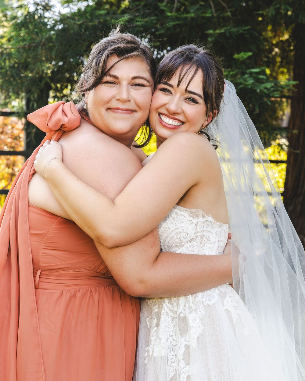 Bride in lace gown and veil shares joyful hug with bridesmaid in coral dress outdoors.