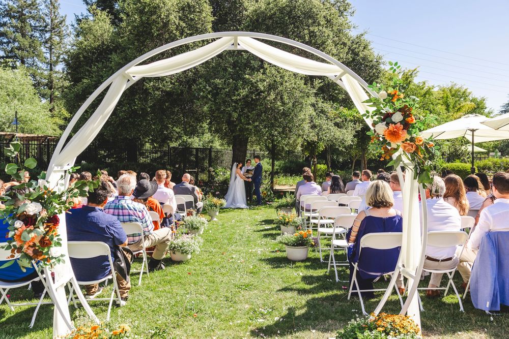 Outdoor wedding ceremony with floral arch, white chairs, and guests seated on sunny lawn surrounded by trees.