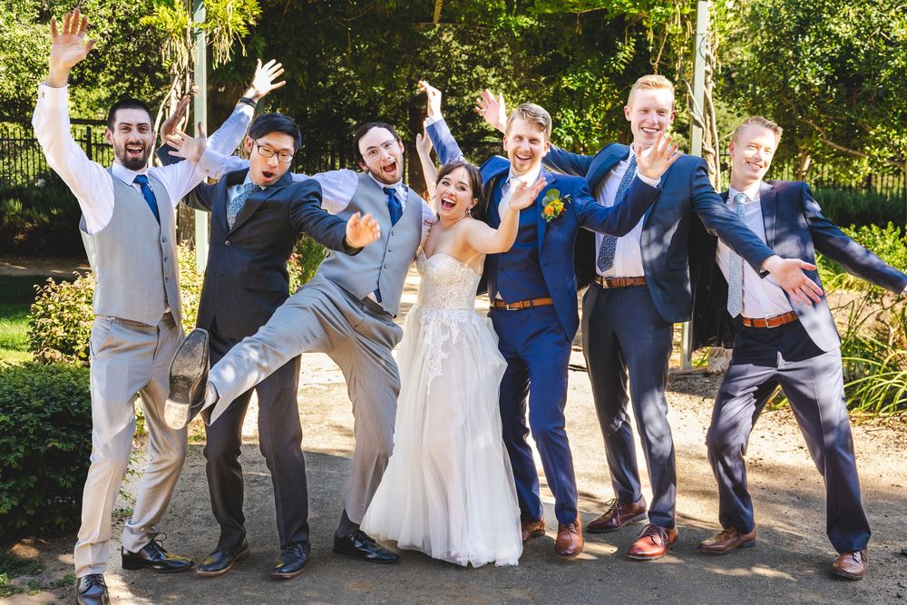 Joyful wedding party posing outdoors with bride in white gown surrounded by groomsmen in navy and gray suits.