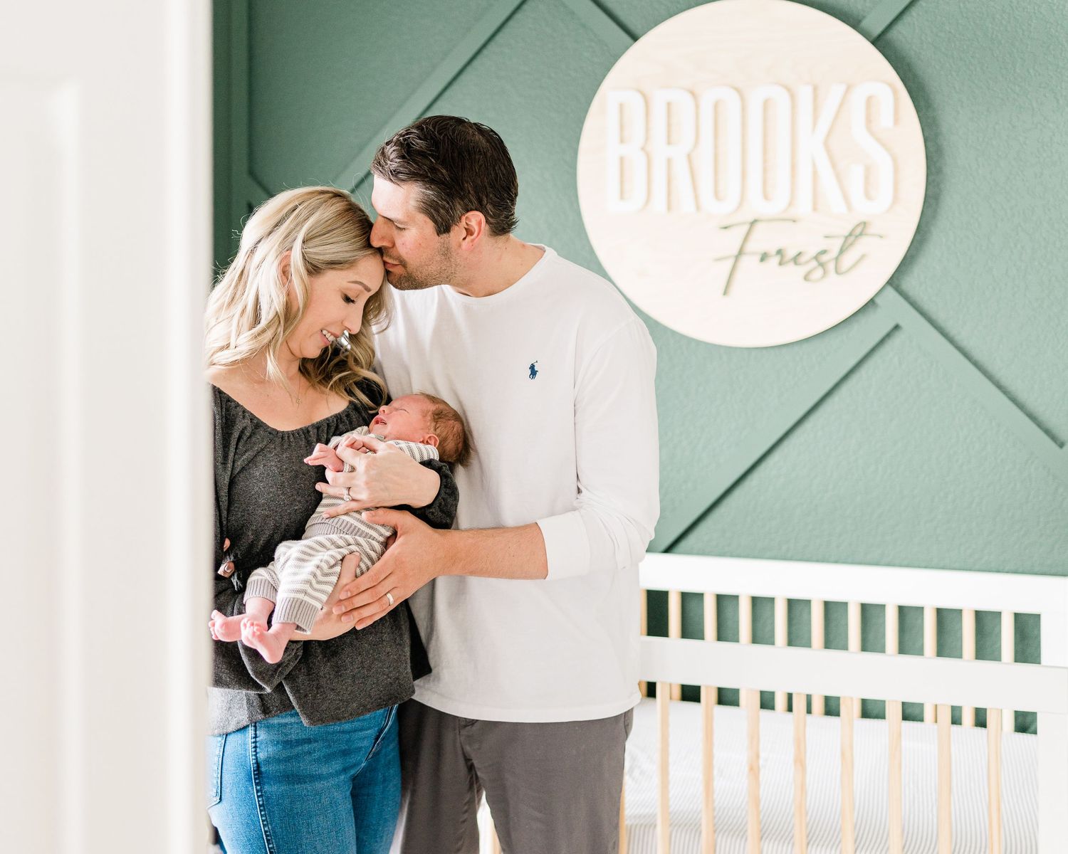 New parents share a tender kiss over their newborn in a nursery with a green accent wall.