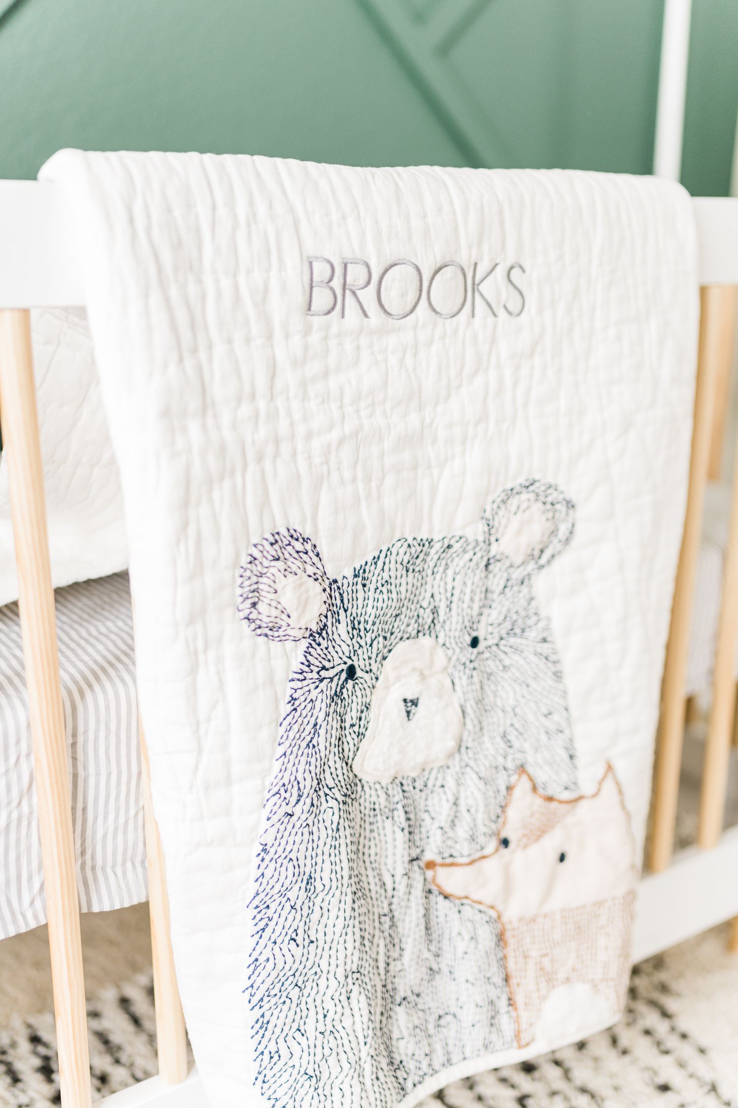 Personalized white quilted baby blanket embroidered with name Brooks and woodland bear design on white crib.
