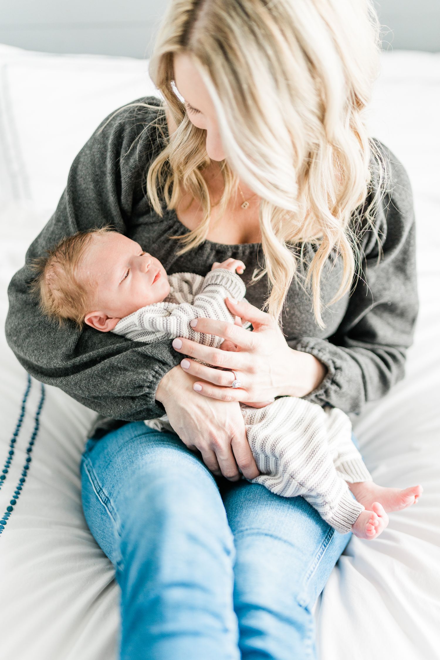 Smiling blonde mother in gray top and jeans tenderly holds sleeping newborn baby in striped outfit on white bed.