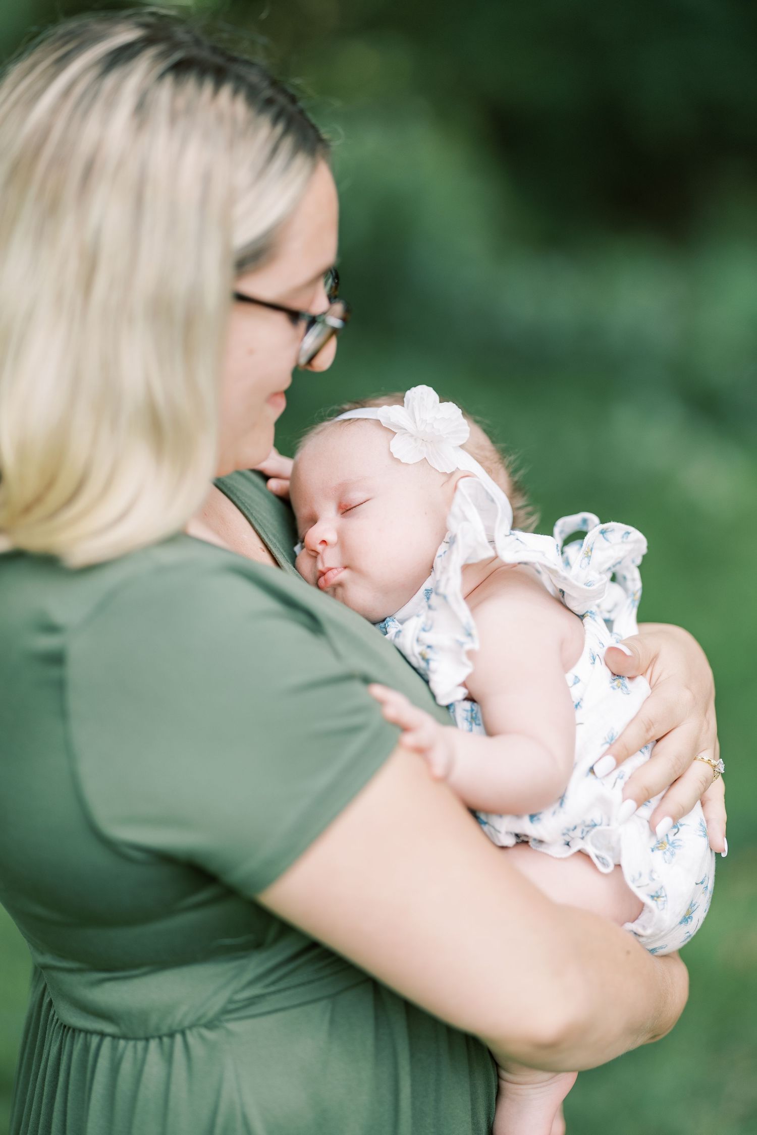 A mother in a green dress tenderly holds her sleeping newborn baby dressed in white outdoors.