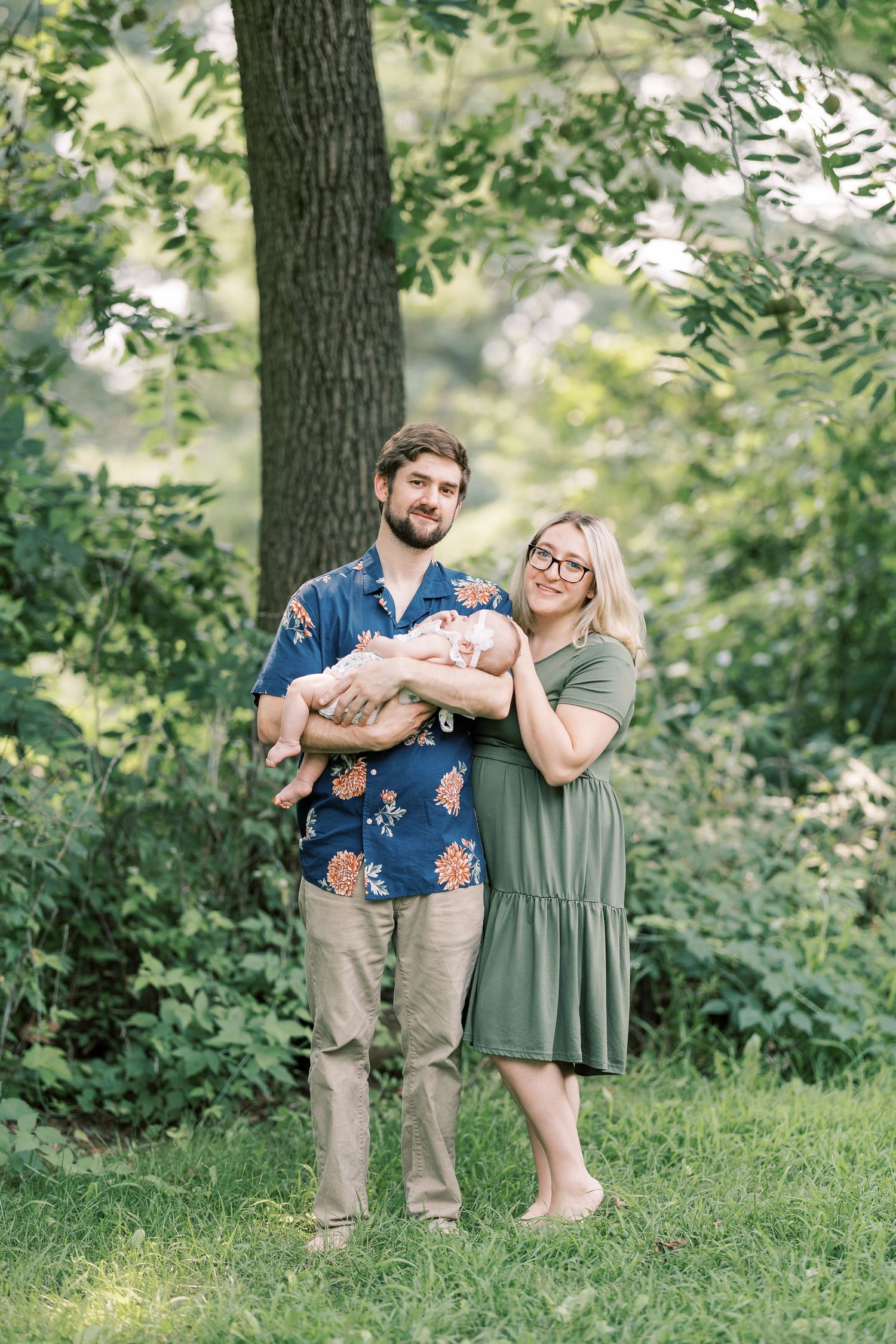 Young family standing outdoors in lush green park, father holding sleeping newborn baby girl.