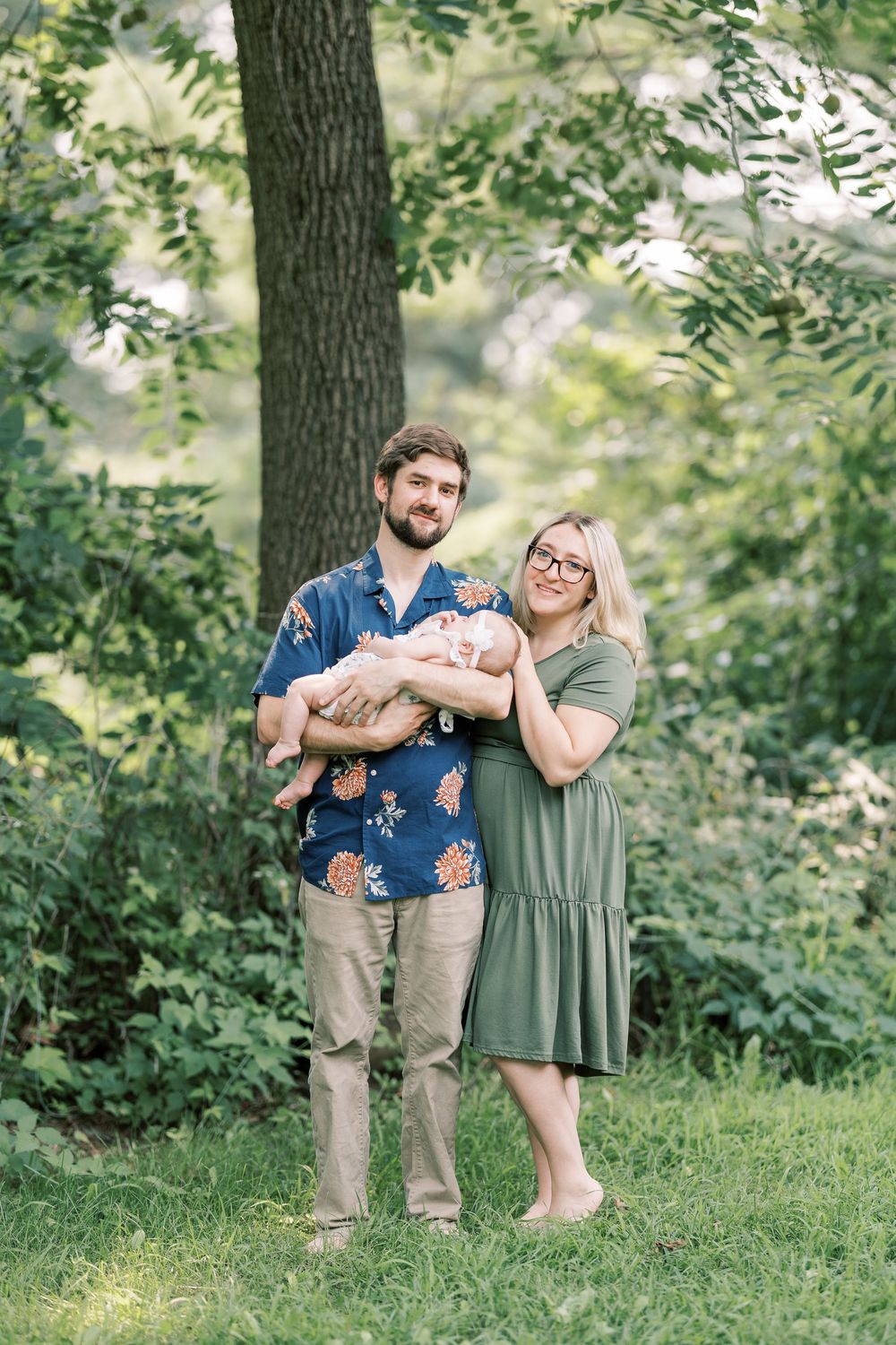 Young family standing outdoors in lush green park, father holding sleeping newborn baby girl.