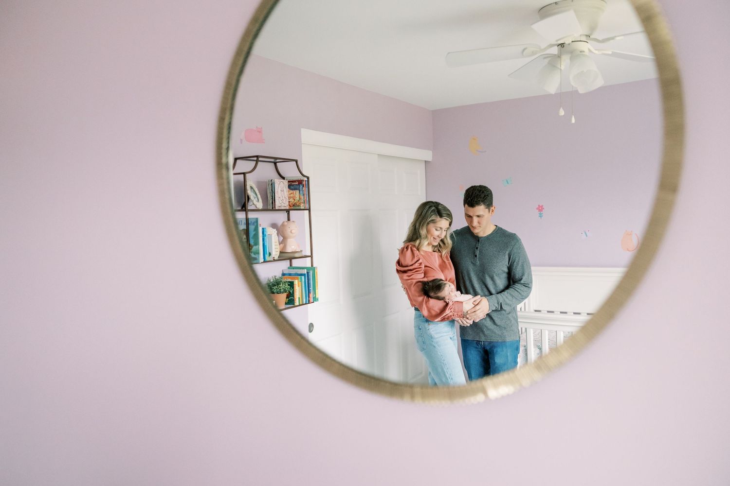 Young couple with newborn baby reflected in round gold-framed nursery mirror.