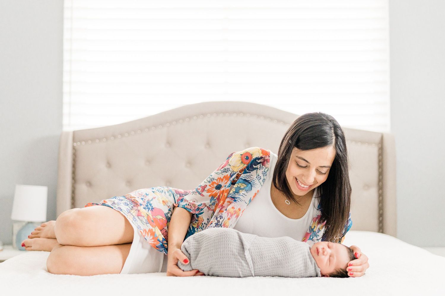 New mother in floral dress lying on bed smiling lovingly at her swaddled newborn baby.