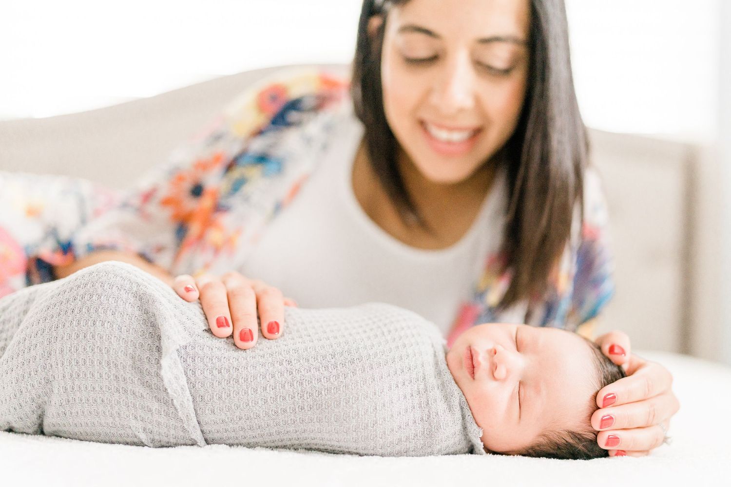 Close-up of sleeping swaddled newborn with smiling mother in floral dress in background.