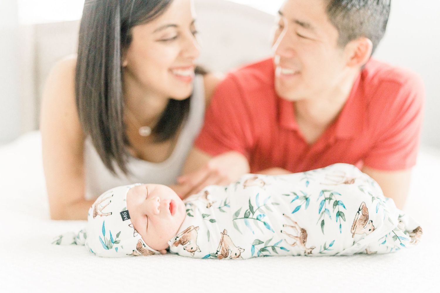 New parents lying on bed smiling at their swaddled newborn baby in floral wrap.