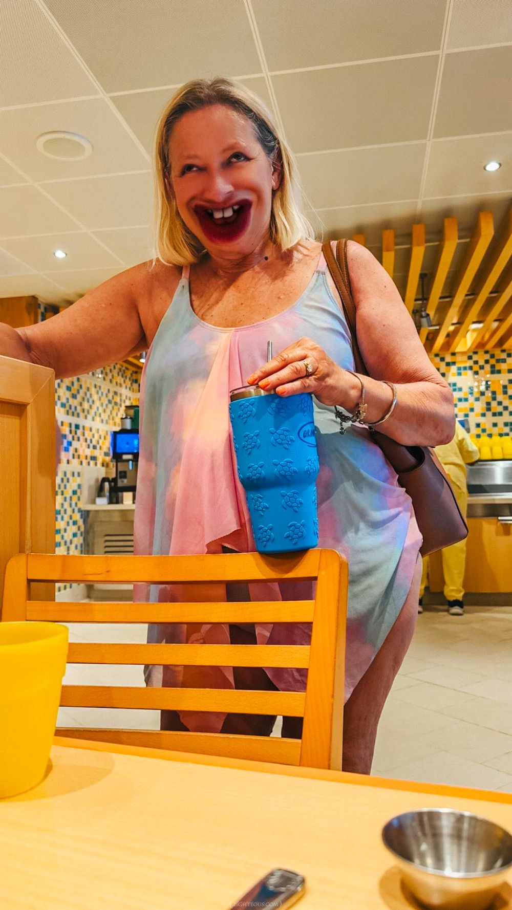 A laughing woman in a colorful dress holds a blue item at a bright yellow dining area on a cruise ship.