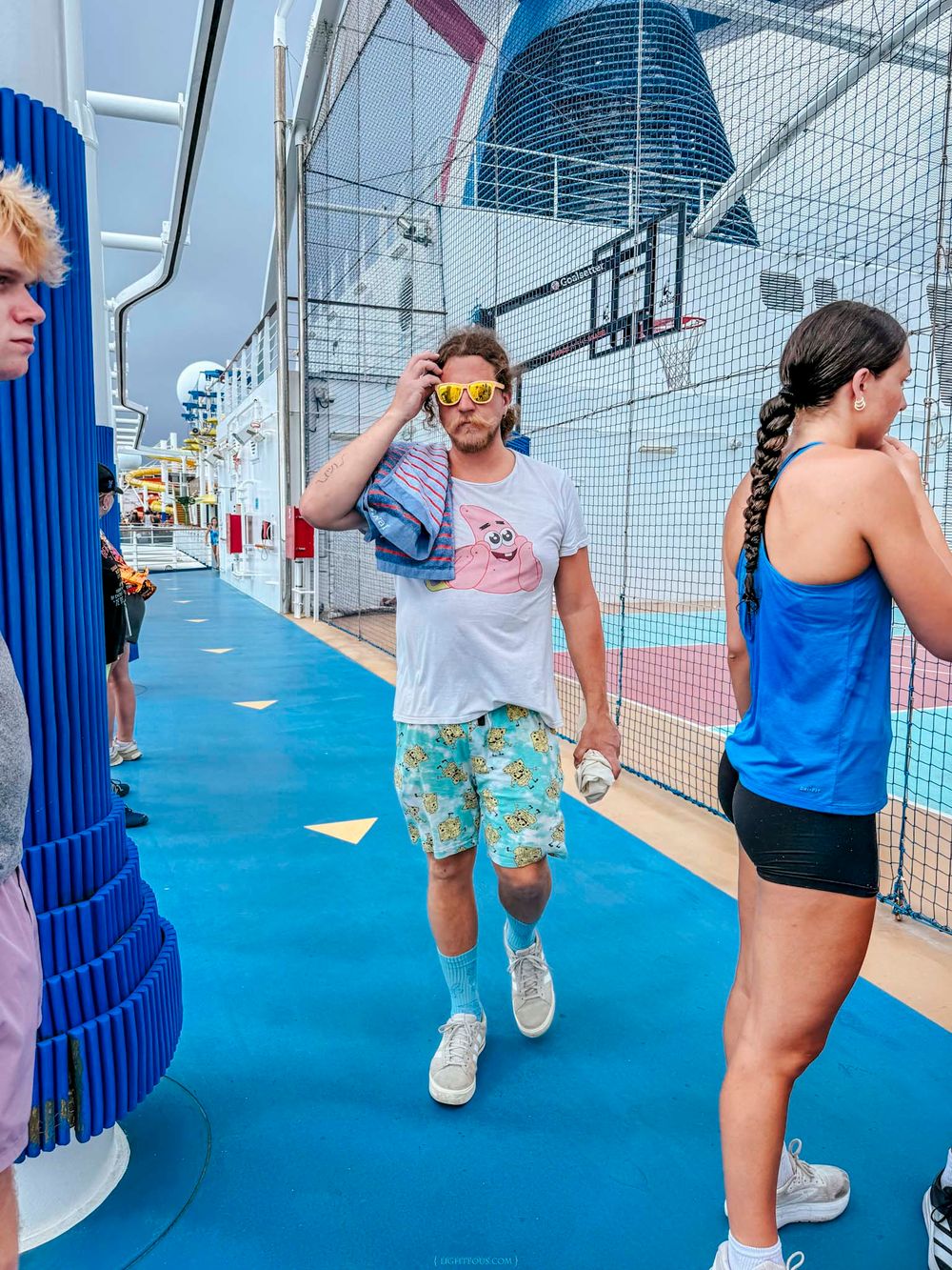 A man in floral shorts and yellow sunglasses walks on a cruise ship sports deck carrying a bag.