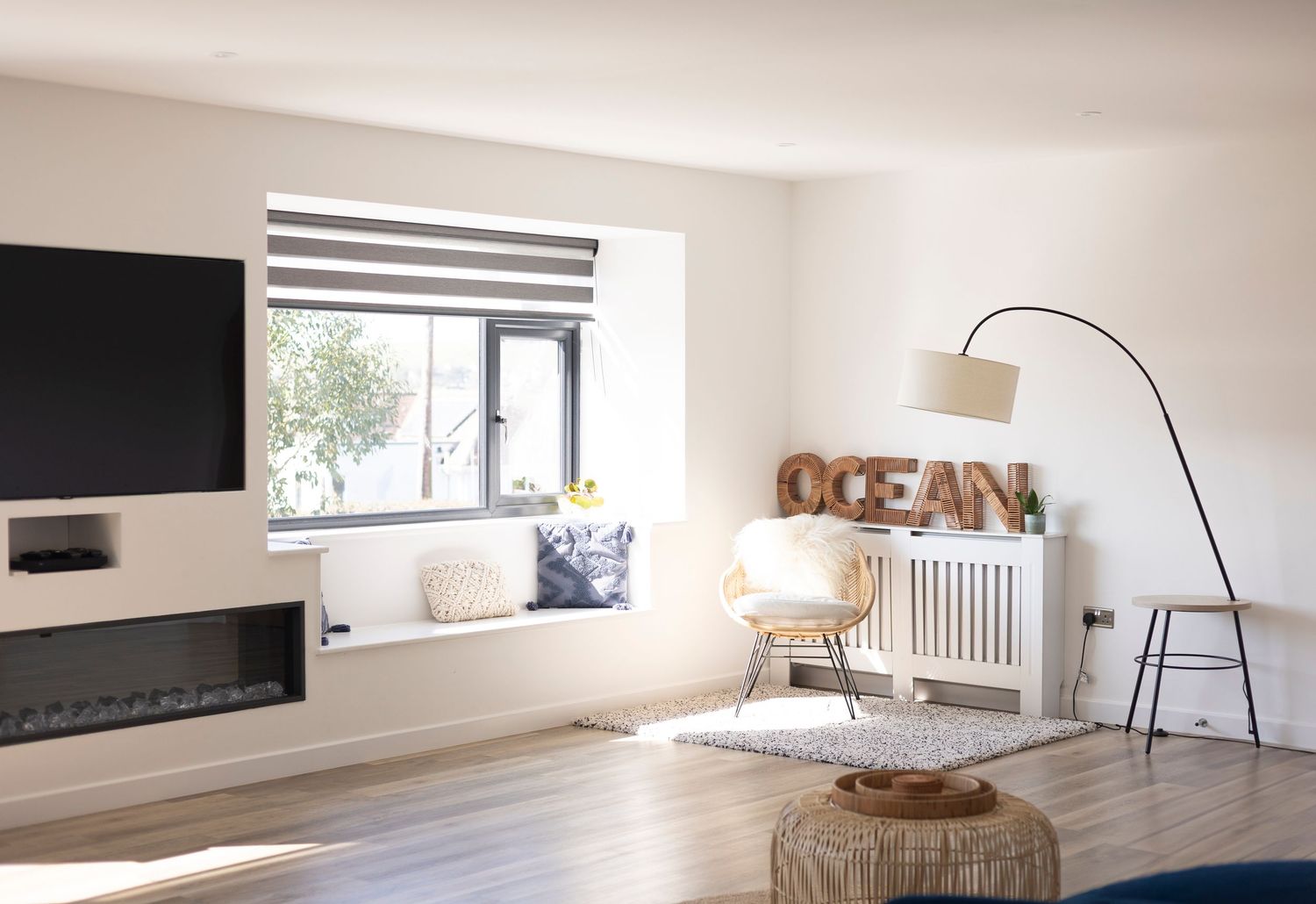 Modern coastal living room with wall-mounted TV, electric fireplace, window seat, and OCEAN sign decor.