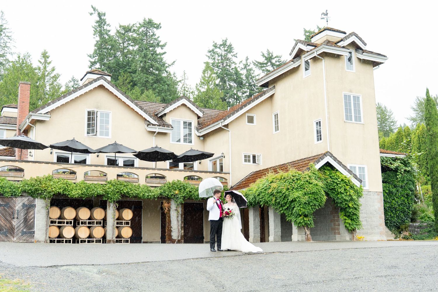 Bride and groom pose in front of a charming winery venue with wine barrels and lush greenery.