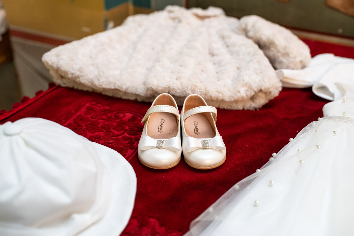 White Mary Jane baptism shoes displayed on red velvet beside a fluffy white fur coat.