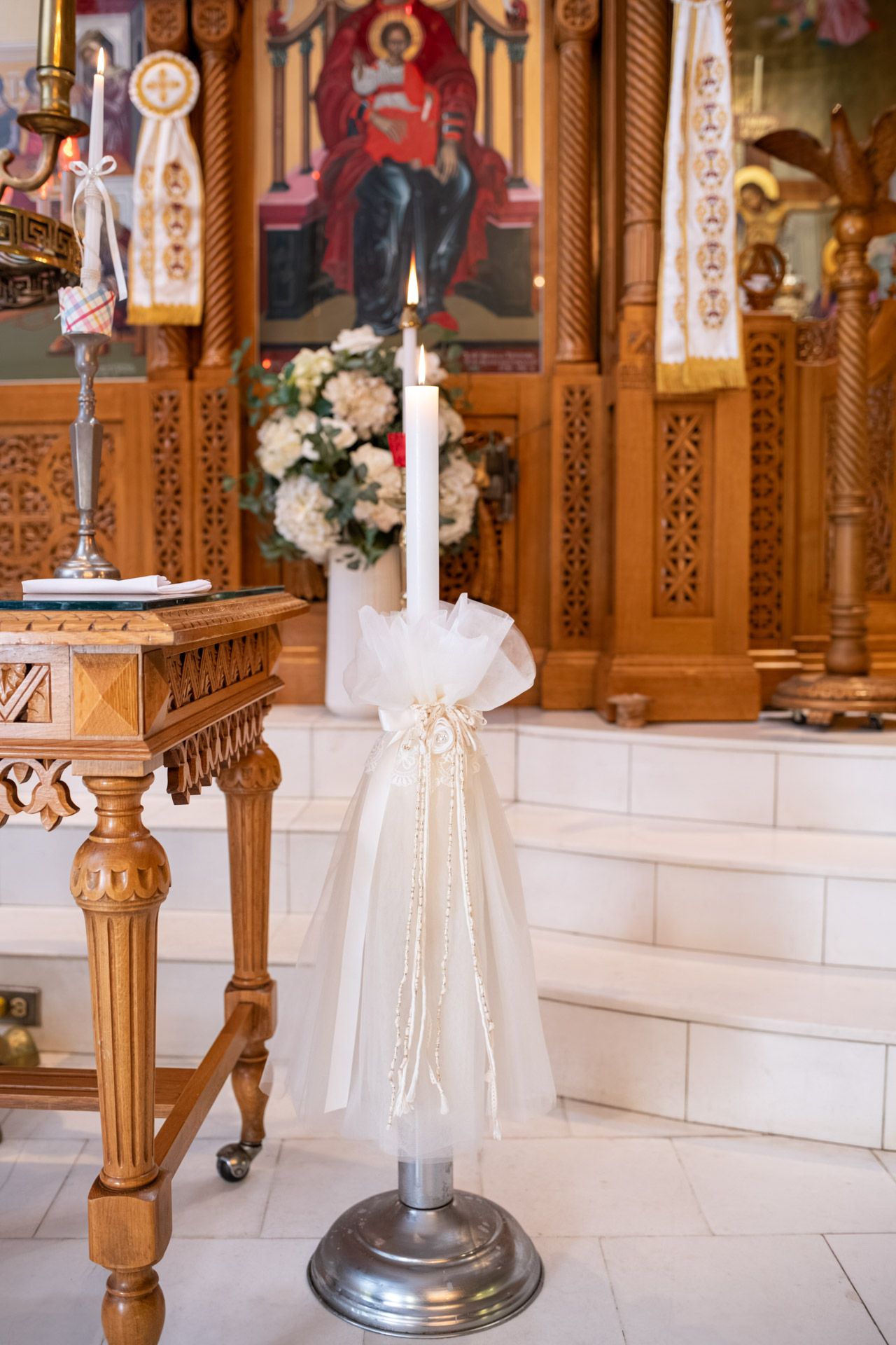 Elegant Orthodox church baptism candle decorated with white tulle, ribbon, and floral arrangement on silver stand.