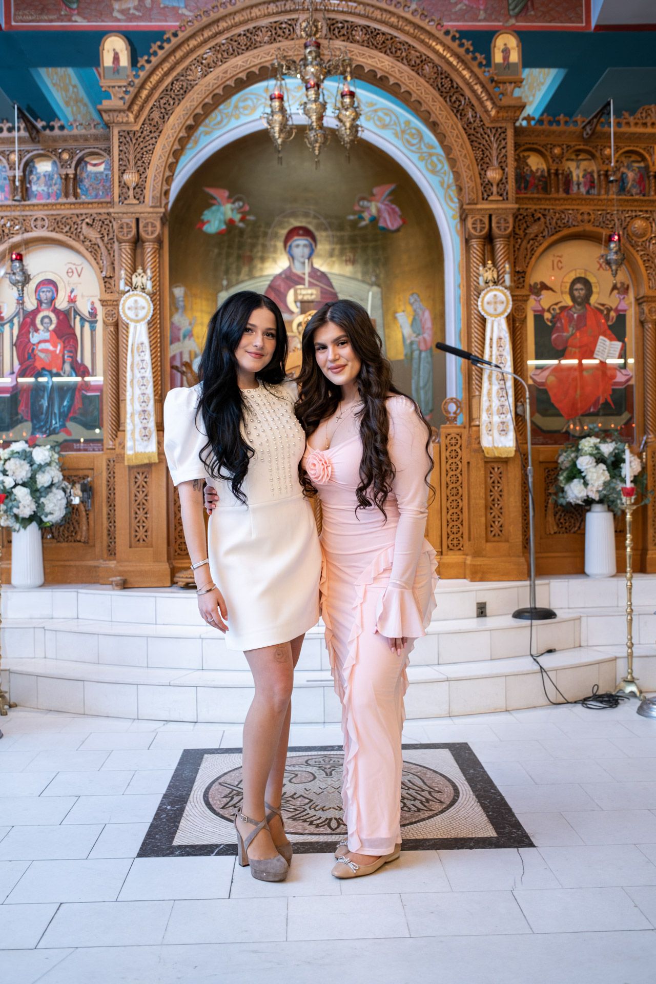 Two young women posing together inside an Orthodox church with ornate iconostasis and religious icons.