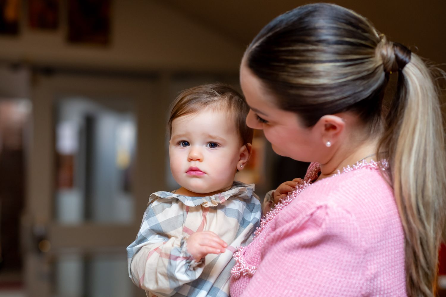 Young mother in pink jacket tenderly holding her baby girl in plaid dress at a christening event.