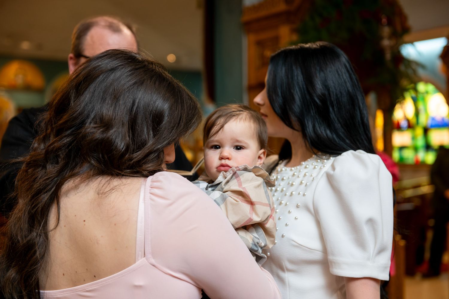 Two women in pink and white dresses admire baby during Orthodox church baptism celebration.