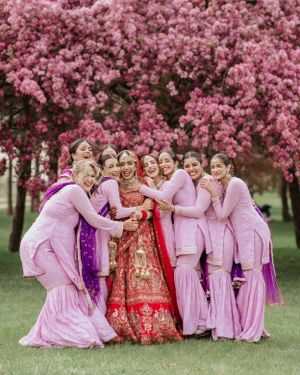 A wedding party in pink traditional attire poses together under blooming cherry blossom trees in a spring garden setting.