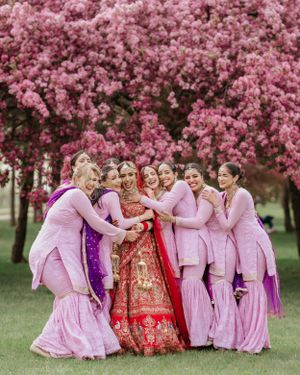 Bridesmaids in pink outfits huddle together with bride in traditional red Indian lehenga under blooming cherry blossoms.