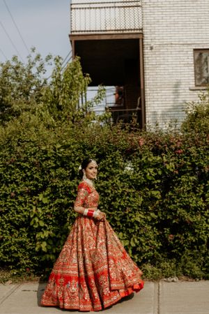 Elegant red and gold embroidered lehenga captured against white brick building and garden setting.