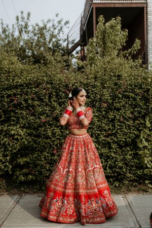 A person wearing an ornate red traditional Indian lehenga with detailed embroidery poses in front of green hedges.