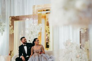 An elegantly dressed couple sits together amid white decor and twinkling lights at a formal celebration.