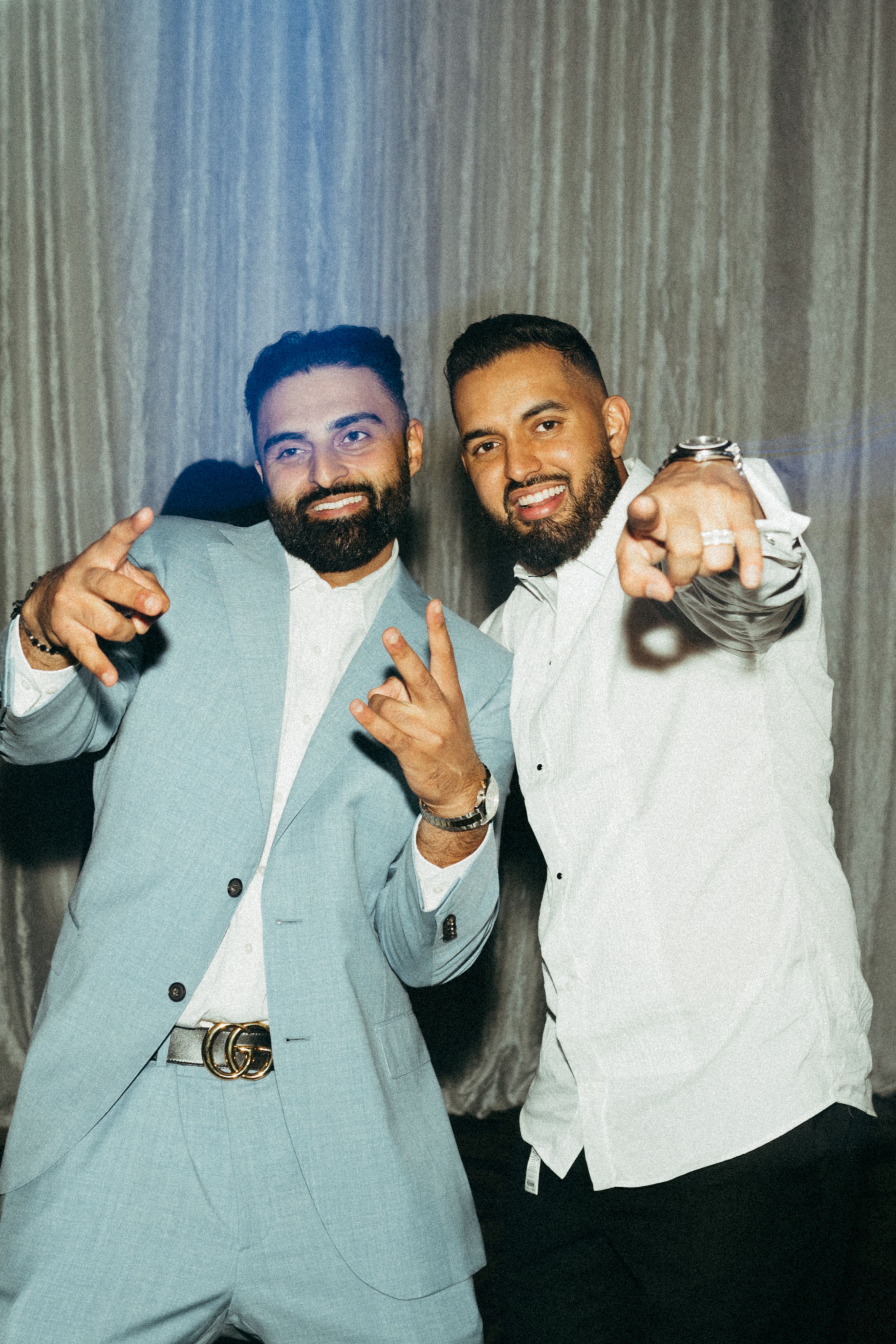 Two friends in coordinating light suits pose together making peace signs at a celebratory event.