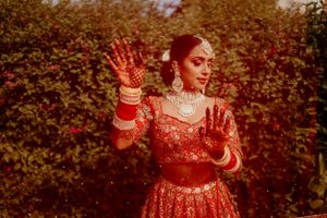 Person in traditional red Indian bridal attire showing decorated henna hands against a natural outdoor backdrop.