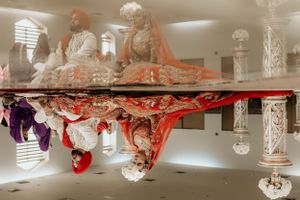 An anamorphic mirror art installation reflects distorted red and white sculptures against a white architectural space.