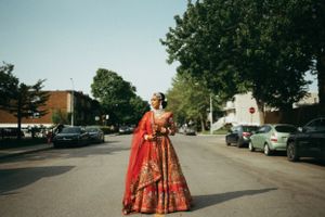 Stunning red bridal lehenga photographed on suburban street with parked cars and trees in background.