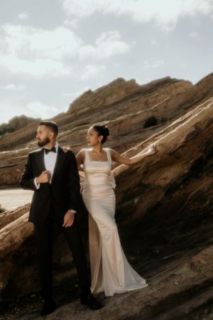 A wedding couple poses among dramatic rocky terrain at sunset, the bride in white and groom in black tuxedo creating contrast.