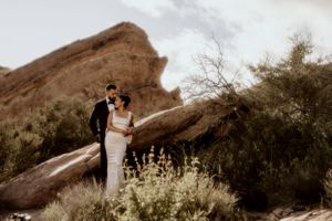 Romantic desert wedding photo with couple posing near large rock formations and desert flora.