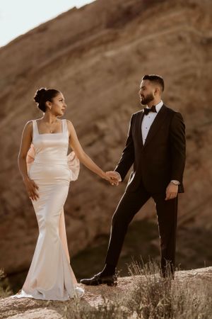 Romantic desert wedding photo of couple in formal attire holding hands against dramatic mountain backdrop at sunset.
