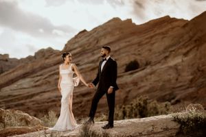 A couple stands together against dramatic red rock formations in formal wedding attire at sunset.