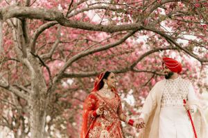 A couple in red and ivory wedding outfits walking together under pink cherry blossom branches.