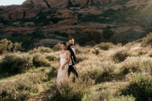 A couple walks through golden grass with majestic red rock cliffs in the background.