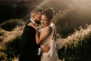 A couple shares an intimate moment in golden sunlight surrounded by wildflowers and rolling hills.