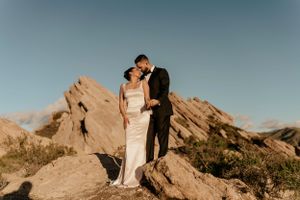 A couple shares an intimate moment atop dramatic rock formations under a blue sky at sunset.
