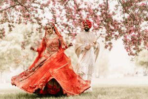 A couple in traditional South Asian wedding attire dance beneath blooming pink cherry blossoms on a misty spring day.