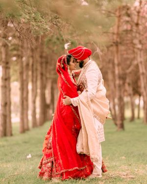 A couple in traditional South Asian wedding attire embrace outdoors among trees, with the bride in a vibrant red lehenga.
