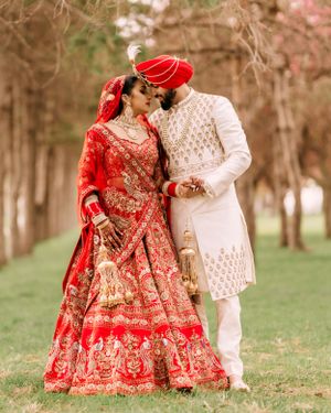 A couple in traditional Indian wedding attire, with the bride in red lehenga and groom in white sherwani, pose outdoors.