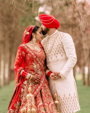 A couple in traditional South Asian wedding attire, with the bride in red and groom in white, share an intimate moment outdoors.