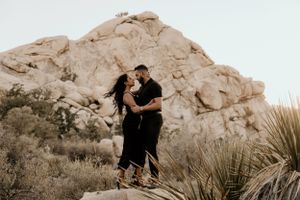 Romantic couple in black outfits pose together against desert rock formations at dusk.